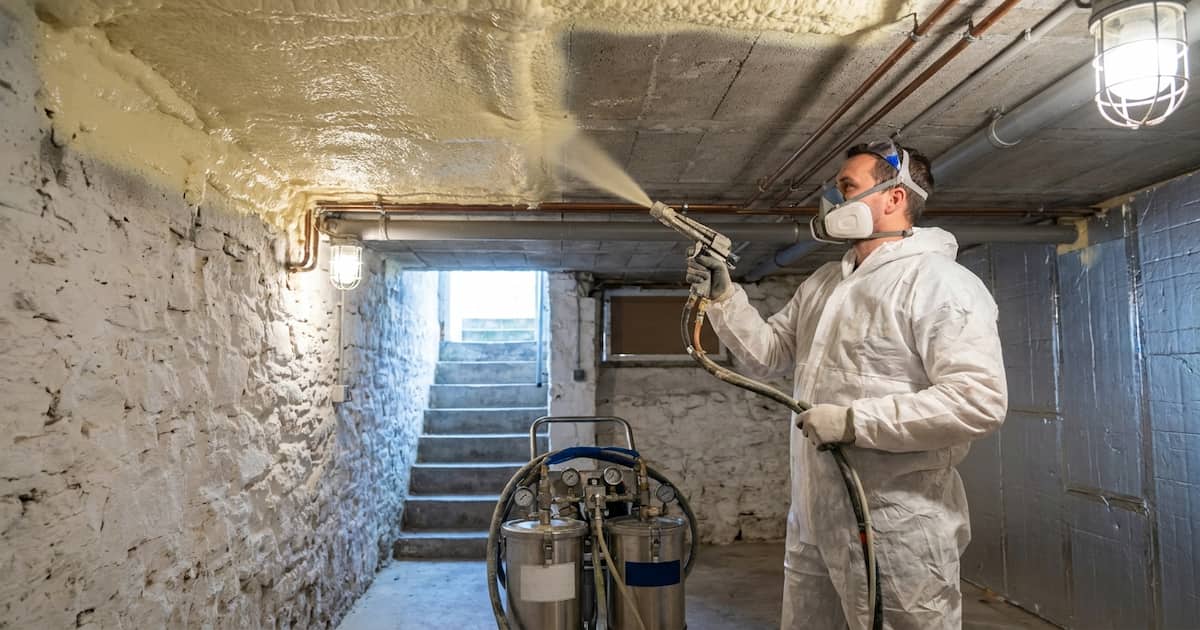 Un artisan en combinaison de protection projette de la mousse polyuréthane sur le plafond en béton brut d'une cave belge, la mousse crème enrobant les canalisations tandis que la moitié non traitée du plafond montre le contraste avec le béton nu