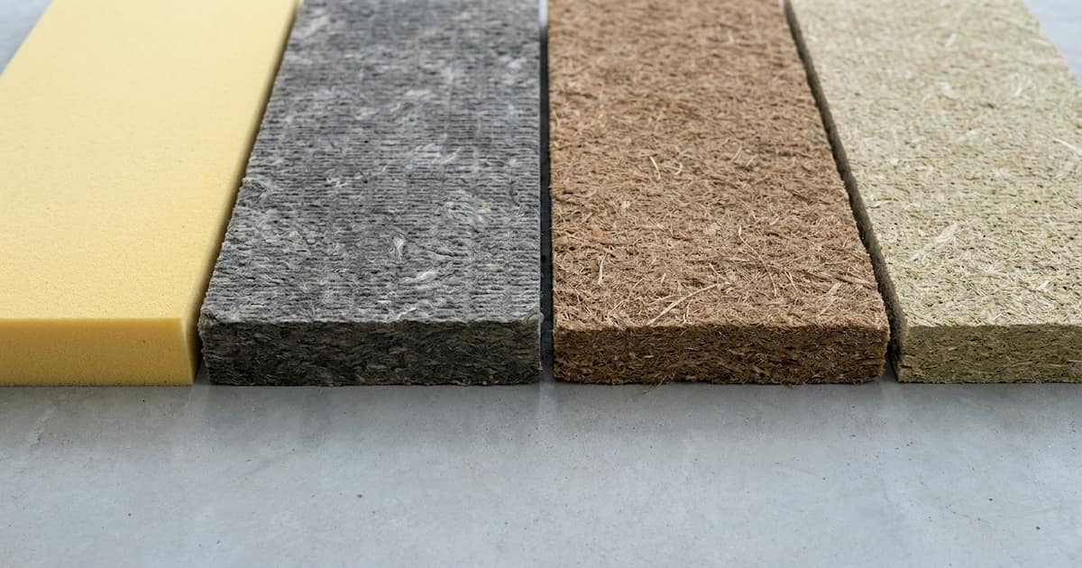 Quatre échantillons d'isolants thermiques vus de dessus sur une surface grise, montrant les textures distinctes du polyuréthane jaune, de la laine de roche grise, de la fibre de bois brune et d'un panneau biosourcé beige-vert.